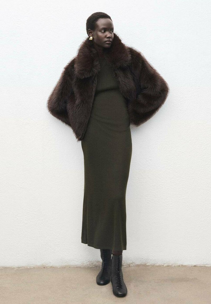 Giacca in pelliccia sintetica marrone con colletto alto sopra un vestito aderente verde scuro. Stivaletti neri con lacci completano l'outfit. Sfondo semplice.