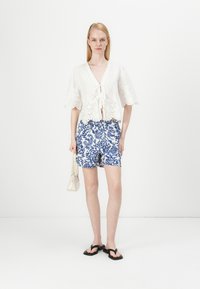 Valge broderie anglaise top, mida kaunistab seotud esiosa, on paaris siniste ja valgete lillemustriga lühikesed püksid ning mustad plätud, käes on väike kreemivärvi käekott.