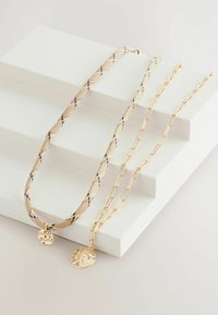 Schichtketten mit einem strukturierten goldenen Anhänger, einer gemusterten Netz- und einer klassischen Gliederkette, alle in Goldtönen auf einem hellen Hintergrund.