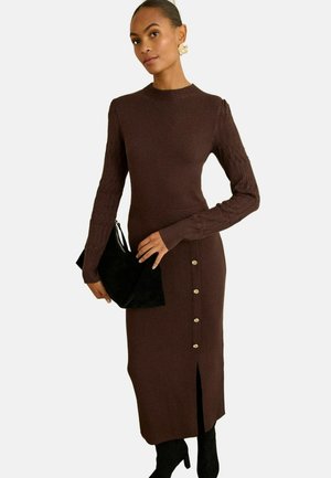 Brun stickad klänning med långa ärmar, ribbad textur, sidoklyfta och detaljer i guldknappar. Kombinerad med en svart clutch.