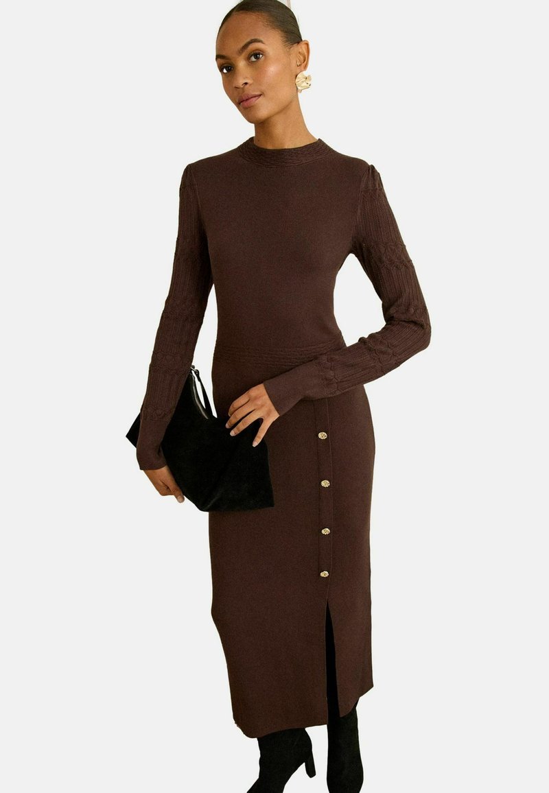 Abito di maglia marrone con maniche lunghe, texture a coste, spacchi laterali e dettagli con bottone dorato. Abbinato a una pochette nera.