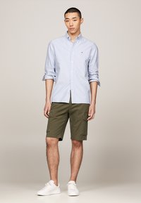Chemise à boutons bleu clair et blanche à rayures, avec les manches retroussées, associée à un short cargo vert olive et des baskets blanches.