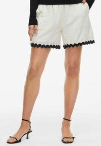 White linen shorts featuring a black scalloped hem and side pockets, paired with black strappy sandals showcasing a thin ankle strap.