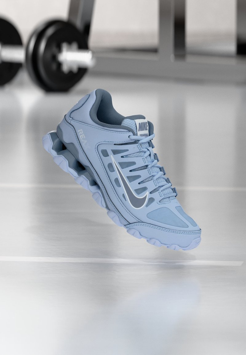 Ανοιχτό μπλε αθλητικό παπούτσι Nike με μαύρο σήμα, λεπτομερές πέλμα, αιωρείται πάνω από ανακλαστικό πάτωμα γυμναστηρίου με θολά βάρη στο φόντο.