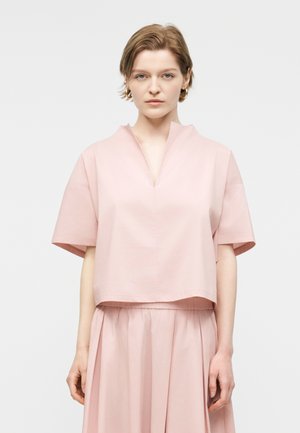 Kvinde med kort lyst brunt hår iført en løstsiddende, lys lyserød kortærmet top og matchende plisseret nederdel, stående mod en ensfarvet baggrund.