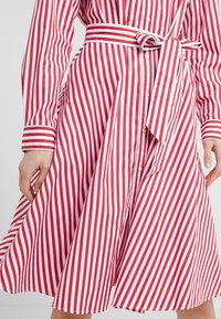 Czerwona i biała sukienka w paski z długimi rękawami, zapinana na guziki z przodu i związywanym paskiem w talii. Materiał wydaje się gładki z opadającym dołem.