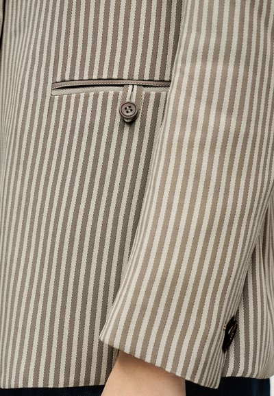 Μανίκι σακακιού με κάθετες ρίγες σε μπεζ και καφέ χρώμα, με λεπτομέρεια τσεπάκι με κουμπί και εμφανές τον καρπό ενός ατόμου κάτω από τη μανσέτα.