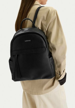 Persona con capelli castani lunghi indossa una giacca beige e porta uno zaino Lasocki nero texturizzato con cerniera e tasca frontale.