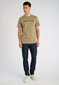 Khaki katoenen t-shirt met "COPENHAGEN" en "LINDBERGH" in donkere letters, gecombineerd met donkerblauwe jeans en witte sneakers.