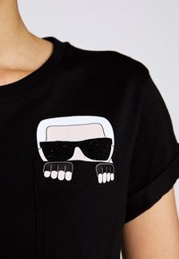 Rysunkowa postać z białymi włosami i dużymi czarnymi okularami przeciwsłonecznymi wygląda zza kieszeni na czarnej koszulce noszonej przez osobę.