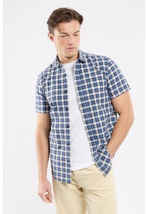 Homme portant une chemise à manches courtes à carreaux bleu et blanc par-dessus un t-shirt blanc et un pantalon beige, avec une main dans la poche, regardant vers le bas à gauche.