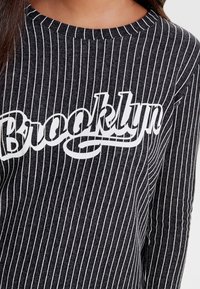 Musta-valkoiset raidalliset pitkähihaiset paita, jossa on pyöreä kaula-aukko ja jossa on näkyvä "Brooklyn" -painatus valkoisena rinnassa.