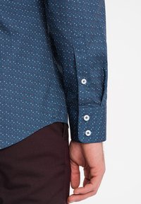 Camicia blu navy con bottoni e motivo a pois rosso e bianco. Presenta un colletto a punta e polsini con bottone. Pantaloni bordeaux scuro.