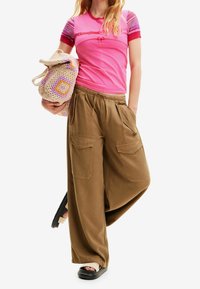 Rosa t-shirt med korta, flerfärgade randiga ärmar, parat med lösa bruna cargobyxor med framfickor. Håller en virkad väska.