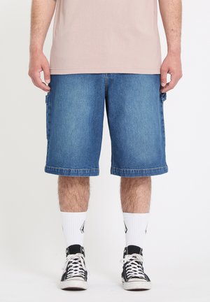 Bas du corps d'une personne portant un short en denim bleu mi-long, des chaussettes blanches montantes avec des logos, et des baskets montantes noires avec des lacets blancs.