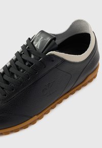 Black leather sneaker with textured finish, cream accents, flat laces, and a gum rubber sole. Features a subtle embossed logo on the side.