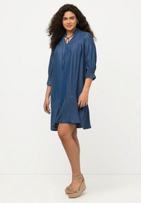 Denimmekko, jossa on väljä malli, 3/4-hihat, avainaukko kaula-aukko, smockatut hihansuut ja kerroksittain muotoiltu helma, keskipainoisessa sinisessä pesussa.