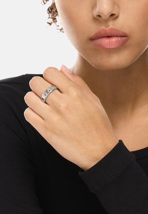 Frau mit hellbrauner Haut, die an ihrer rechten Hand einen silbernen Ring mit einem doppelten 'C'-Logo trägt und ein schwarzes Langarmshirt anhat.