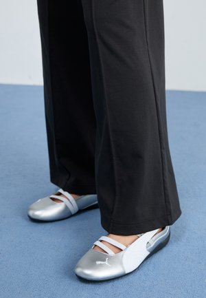 Scarpe piatte argento e bianche con cinturini incrociati e dettagli testurizzati, abbinate a pantaloni neri larghi. Ambientate su un pavimento blu.