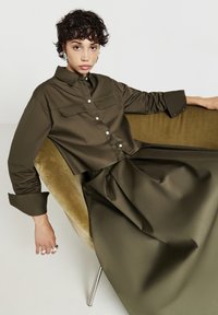 Camicetta verde oliva a maniche corte con abbottonatura e due tasche sul petto, abbinata a una lunga gonna fluida coordinata. Tessuto dalla texture liscia.