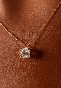 Gouden ketting met ronde, heldere edelsteen in een vierklauwzetting, die contrasterend afsteekt tegen een donkere huidtextuur.