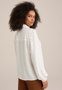 Blusa bianca con un delicato motivo a pois dorati, maniche lunghe a sbuffo e colletto con bottoni. Il tessuto strutturato offre un aspetto leggero e arioso.