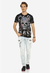 Schwarzes Grafik-T-Shirt mit Schädelfmotiven und Text, kombiniert mit hellblauen Jeans mit Seitentaschen und strukturiertem Muster sowie schwarzen Schuhen.