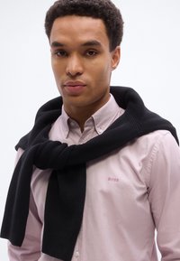 Νέος άνδρας με ανοιχτό ροζ πουκάμισο με κουμπιά και λογότυπο "BOSS", φοράει ένα μαύρο πουλόβερ δεμένο γύρω από τους ώμους του μπροστά σε απλή φόντο.