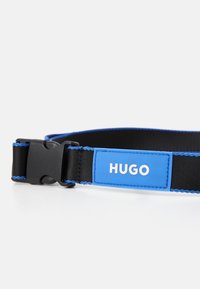 Cinturón negro con acentos azules y una hebilla de plástico. Presenta una etiqueta rectangular azul con "HUGO" en texto blanco y bordes cosidos.