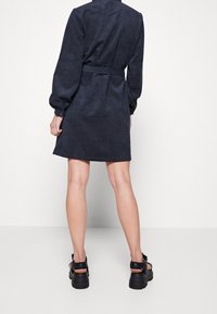 Abito camicia in corduroy blu navy con maniche lunghe a palloncino, vita con cintura e lunghezza fino a metà coscia. Sono indossate sandali neri chunky.