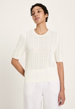 Woman wearing a white short-sleeve knit top with a chevron lace pattern and white linen pants against a plain background.