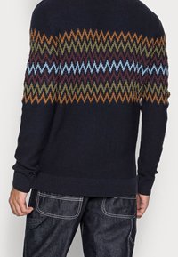 Pull en maille foncé avec un motif en zigzag orange, vert, bordeaux et bleu clair sur le haut du dos et les manches. Poignets côtelés.