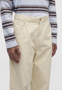 Homme portant un pantalon beige et une chemise à manches longues avec des rayures horizontales marron, blanc et bleu clair, debout les mains détendues.