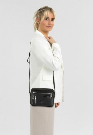 Sac bandoulière noir en matériau texturé, doté de plusieurs poches zippées et d'accents en métal argenté, porté sur un blazer blanc.