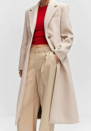 Persona che indossa un lungo cappotto beige, top aderente rosso e pantaloni beige a righe larghe a vita alta con la mano in tasca.