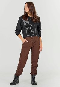 Felpa nera corta con testo e numeri grigi. Pantaloni marroni leggeri e testurizzati con chiusura a due bottoni e tasche. Stivali neri tipo combattimento.