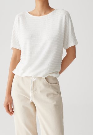 Femme portant un haut blanc texturé à manches courtes légèrement rentré dans un pantalon beige clair à cordon, debout devant un fond uni.