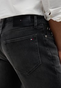 Κοντινό ενός ατόμου που φοράει μαύρα τζιν Tommy Hilfiger με το λογότυπο της μάρκας στην πίσω τσέπη και δερμάτινη ετικέτα στη μέση.