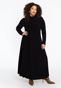 Robe maxi noire à manches longues avec un col montant et un design drapé. Une paire de bottines à talons aiguilles à imprimé léopard et à bout pointu.