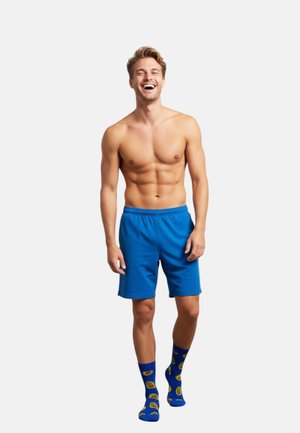 Blaue Baumwollshorts mit elastischem Bund, kombiniert mit blauen Socken mit gelbem Avocado-Muster. Modell ist oberkörperfrei und lächelt.