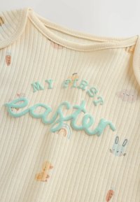 Body color crema con textura acanalada, con gráficos en tonos pastel de patitos, conejitos y arcoíris; "Mi primer Easter" en letras texturizadas color verde azulado.