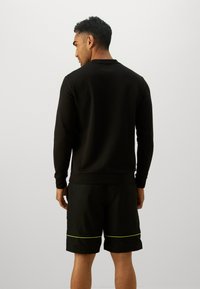 Svart sweatshirt med lange ermer og ribbestrikkede mansjetter, kombinert med svarte shorts som har en neongrønn horisontal stripe.