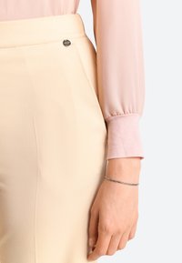 Blouse rose clair à manches longues associée à un pantalon crème comportant un détail de bouton argenté à la taille et une texture lisse.