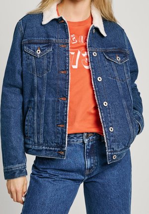 Vrouw die een blauwe denim sherpa-jas draagt over een oranje t-shirt met print, gecombineerd met blauwe denim jeans, staand met één hand in de zak.