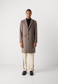 Abrigo de cuadros marrón con un diseño de doble botonadura, cuello con muesca, dos bolsillos frontales y tres botones, que se lleva sobre un cuello de tortuga negro y pantalones beige.