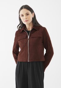 Hnědá semišová bunda s krátkým střihem, dvěma předními kapsami a stříbrným zipem. Stylizováno s černými plisovanými kalhotami.