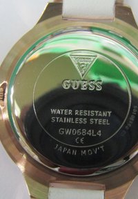 Arrière de montre rond en acier inoxydable avec une surface brillante. Le texte gravé inclut "Guess," "Résistant à l'eau," et "Mouvement Japon," avec un raccord de bracelet blanc.