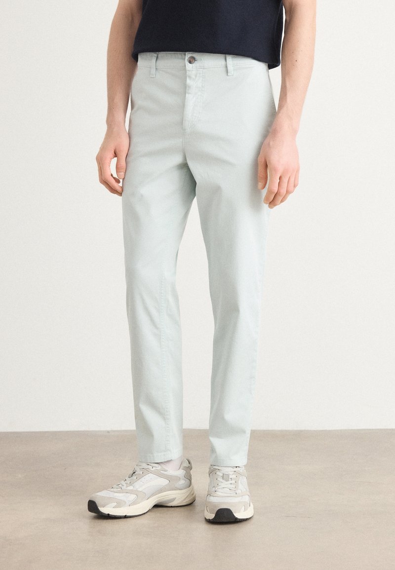 Homme portant un pantalon droit gris clair, des baskets blanches et grises, et un haut noir sur un fond uni.