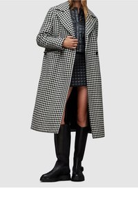 Hahnentritt-Mantel in Schwarz und Weiß mit langer Silhouette, breitem Revers und Seitenschlitzen, getragen über einem gesteppte schwarzen Kleid und kniehohen Stiefeln.