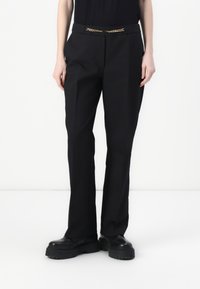 Zwarte broek met een getailleerde pasvorm, voorzien van een gouden kettingaccent bij de taille en een gladde textuur, gecombineerd met dikke zwarte schoenen.